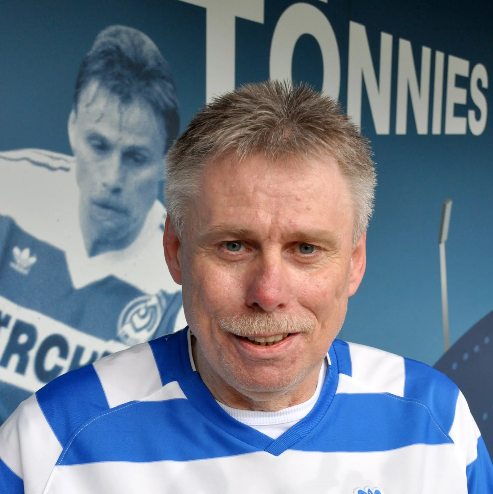 Michael Tönnies lächelt im blau-weißen MSV-Trikot vor einer Wand mit seinem Namen und einem Spielerfoto aus den 1990er-Jahren