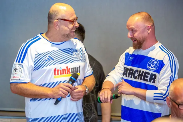 Zwei Männer in MSV-Trikots lachen sich an, links im aktuellen Heimtrikot mit Mikrofon, rechts im Retro-Trikot mit Kärcher-Schriftzug