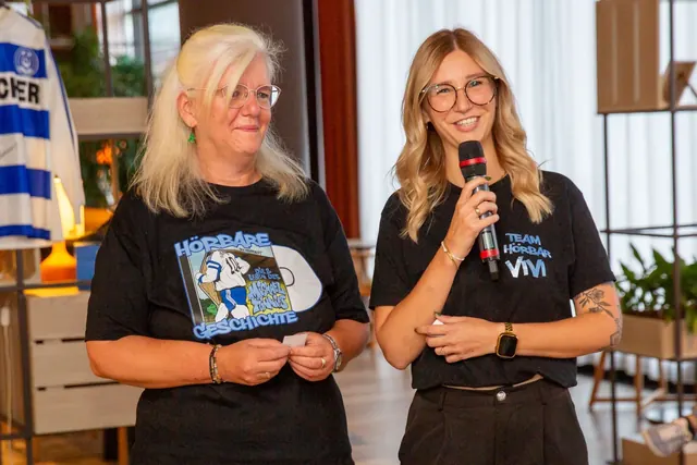 Zwei Frauen im schwarzen T-Shirt: die jüngere mit dem Aufdruck „Team Hörbar Vivi“ spricht ins Mikrofon, die ältere trägt ein T-Shirt „Hörbare Geschichte“