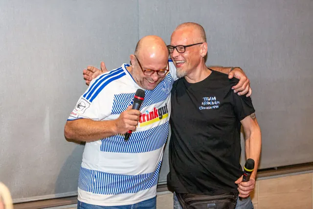Zwei Männer umarmen sich lachend, der rechte trägt ein T-Shirt mit dem Aufdruck „Team Hörbar Christoph“