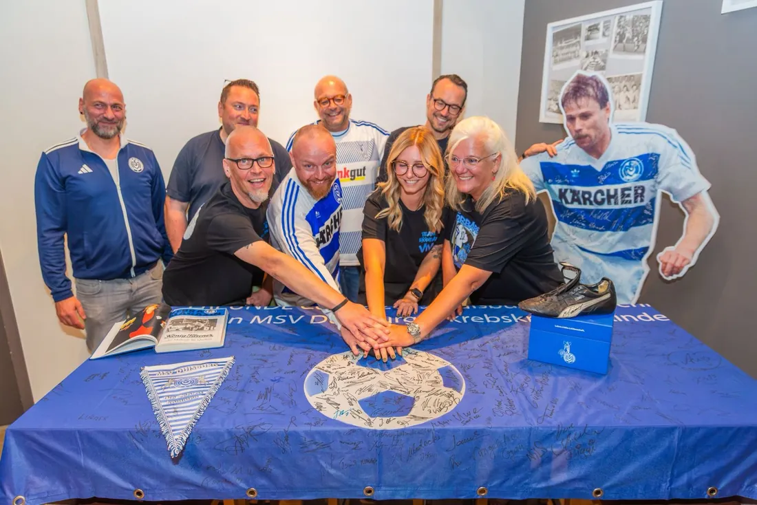 Acht Menschen legen ihre Hände auf ein blaues, vielfach signiertes MSV-Fahnentuch, daneben ein Aufsteller von Michael Tönnies im Kärcher-Trikot