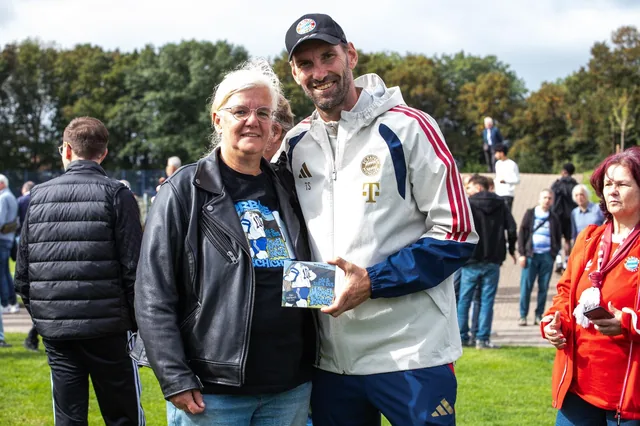 Tom Starke in FC-Bayern-Trainingsjacke hält lächelnd die Hörbuch-CD, neben ihm eine Frau mit grauen Haaren in Lederjacke und Hörbuch-T-Shirt, im Hintergrund Publikum auf einem Rasenplatz